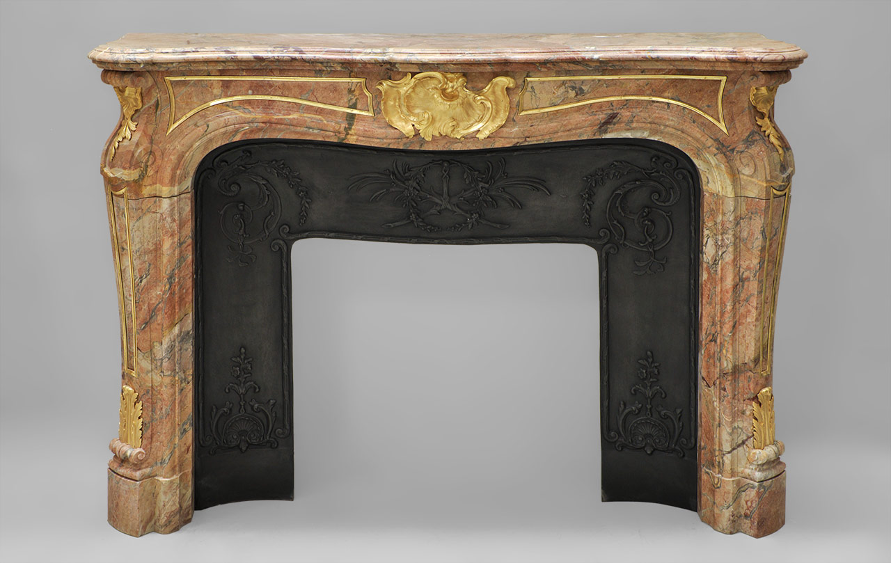 “Comtesse de Vintimille” is a rare Sarrancolin Fantastico marble fireplace mantel with gilded bronze ornaments. This Louis XV style fireplace is shaped with curved contours.
The bronze decoration is both light and opulent. Light thanks to the delicate golden inlays which bring out the panels on the cornice and the legs, but opulent due to the Rocaille ornaments like the central asymmetrical shell which features undulations and curves. The base of the legs is decorated with foliages, while the tops are decorated with an inverted shell.
This fireplace comes with a cast iron insert. The ornaments are gilded.